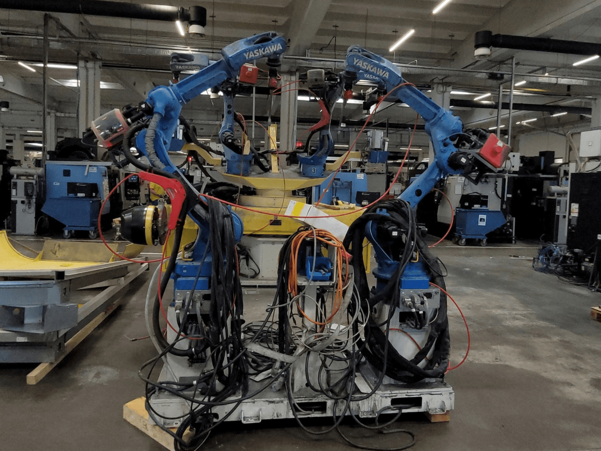 Cellule de soudage robotisée Yaskawa RWC L1_OS 15330, vue de face, équipée de deux robots bleus dotés de nombreux câbles et d'outils de soudage.
