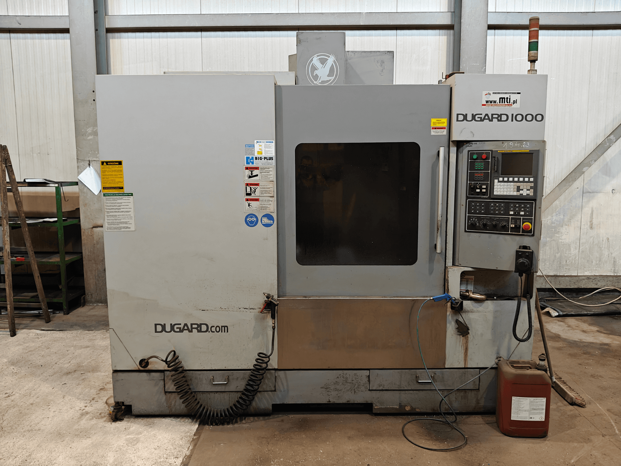 Machine CNC Dugard 1000, vue de face, montrant le panneau de commande, les étiquettes de sécurité et le réservoir d'huile, dans un atelier.