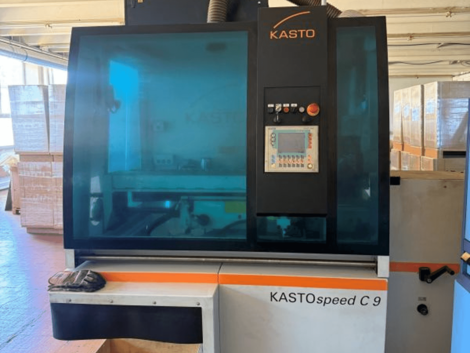Vue de face de la scie circulaire à métaux KASTO KASTOspeed C 9, avec son panneau de commande et sa vitre de protection, et des caisses en bois en arrière-plan.