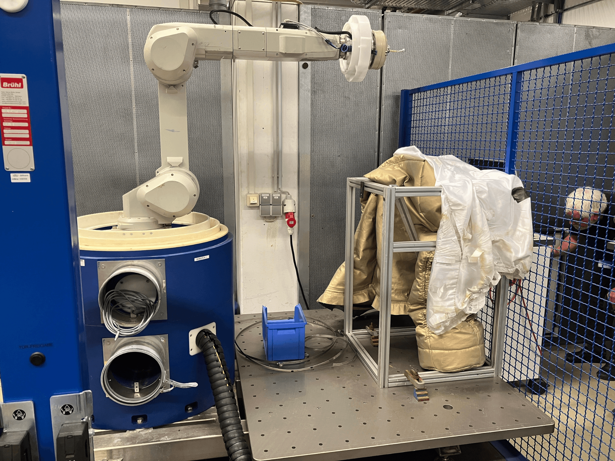 Bras robotique près d'une chambre bleue, tenant un costume blanc et or sur un cadre métallique, vue de côté sur fond de filet de sécurité.