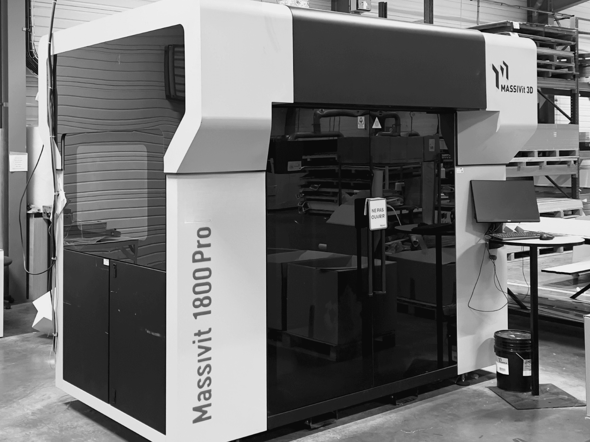 Imprimante 3D en plastique MASSIVIT 1800 PRO dans un atelier, vue de face, avec un poste de travail informatique et un panneau de commande visibles.
