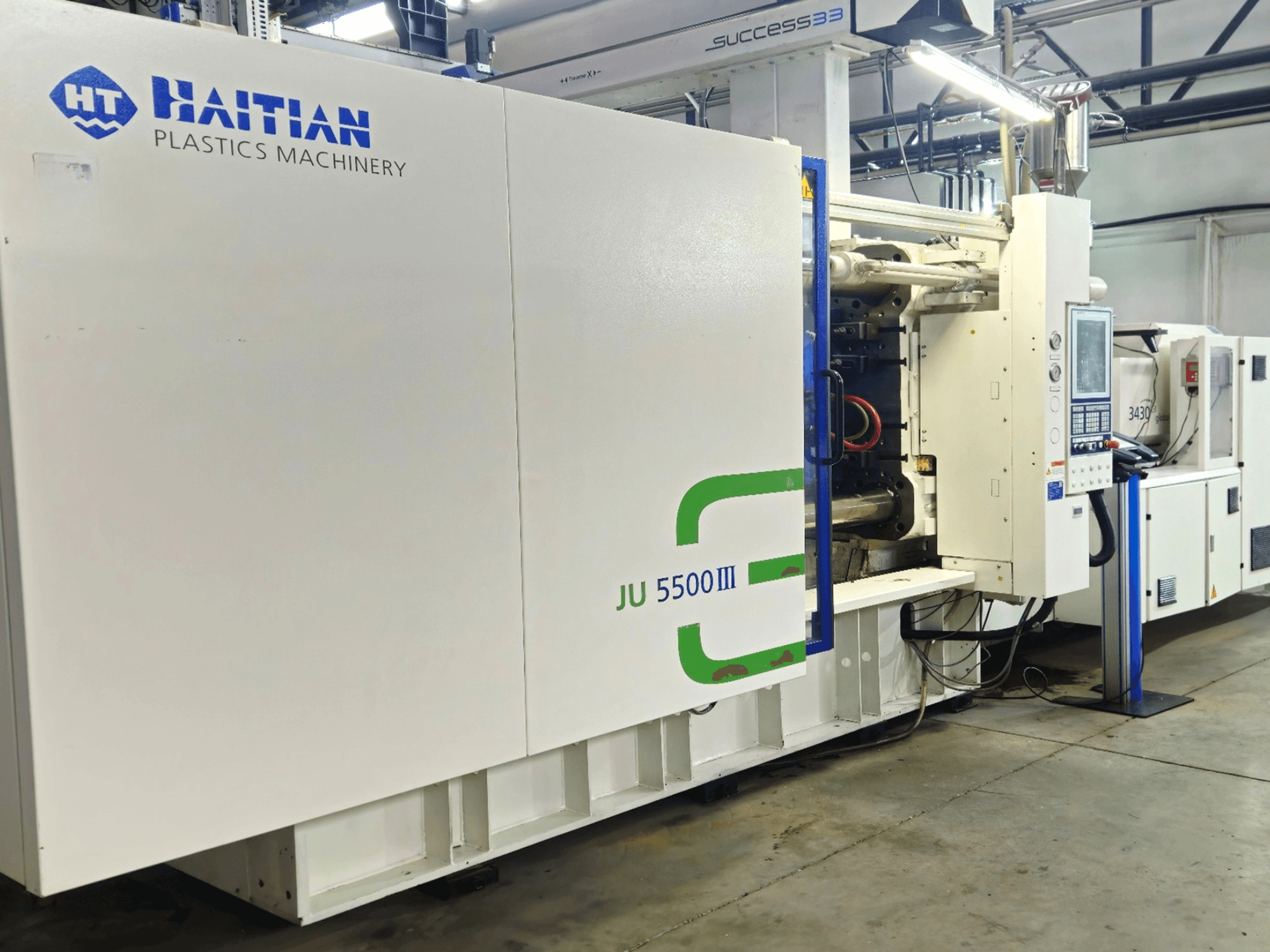 Machine haïtienne JU 5500III pour matières plastiques dans une usine, vue de face montrant le panneau de commande et la zone de production.