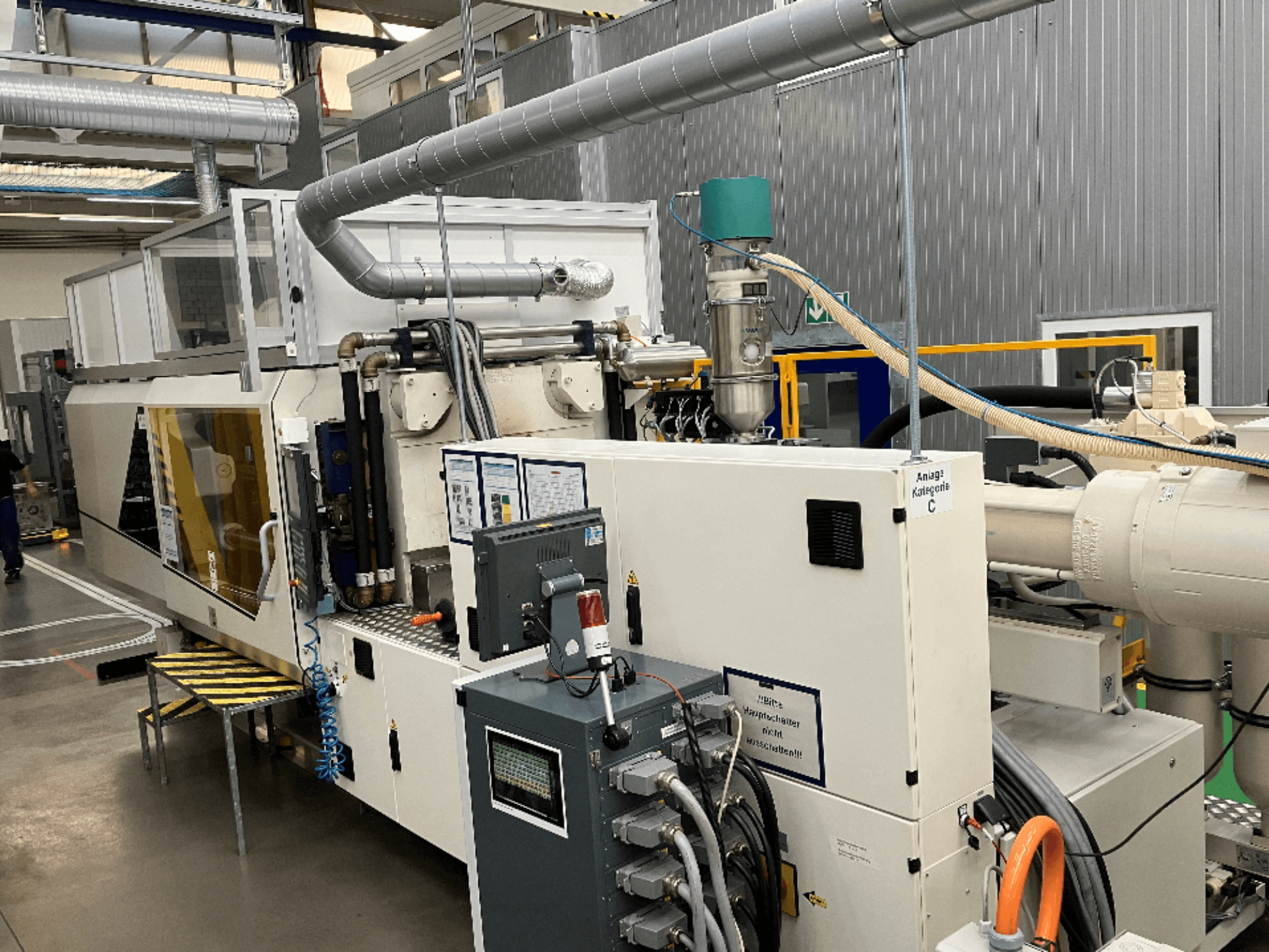 Machine de moulage par injection, vue de face, avec étiquette de marque, panneau de commande et tuyaux visibles dans une usine.