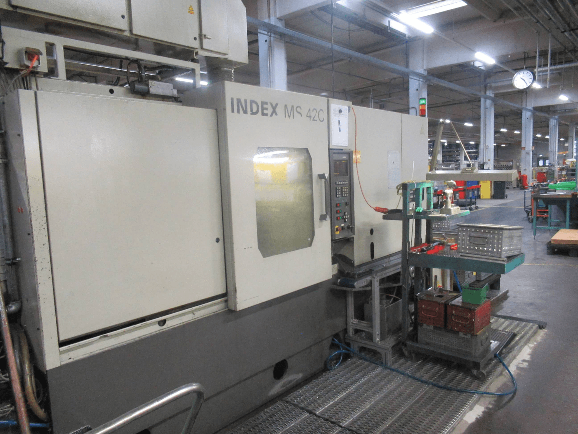 Machine CNC INDEX MS 42C dans un atelier, vue latérale montrant le panneau de commande et le rangement des outils sur un chariot à roulettes.