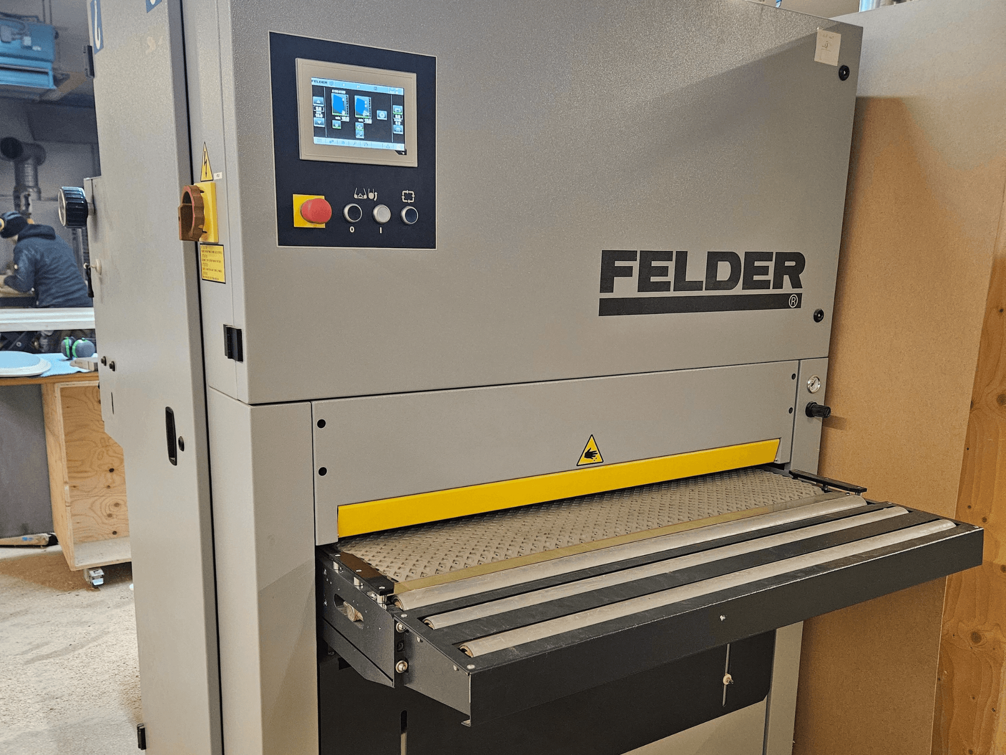 Machine à travailler le bois FELDER vue de face, comportant un panneau de commande numérique et une table d'alimentation jaune avec des avertissements de sécurité.