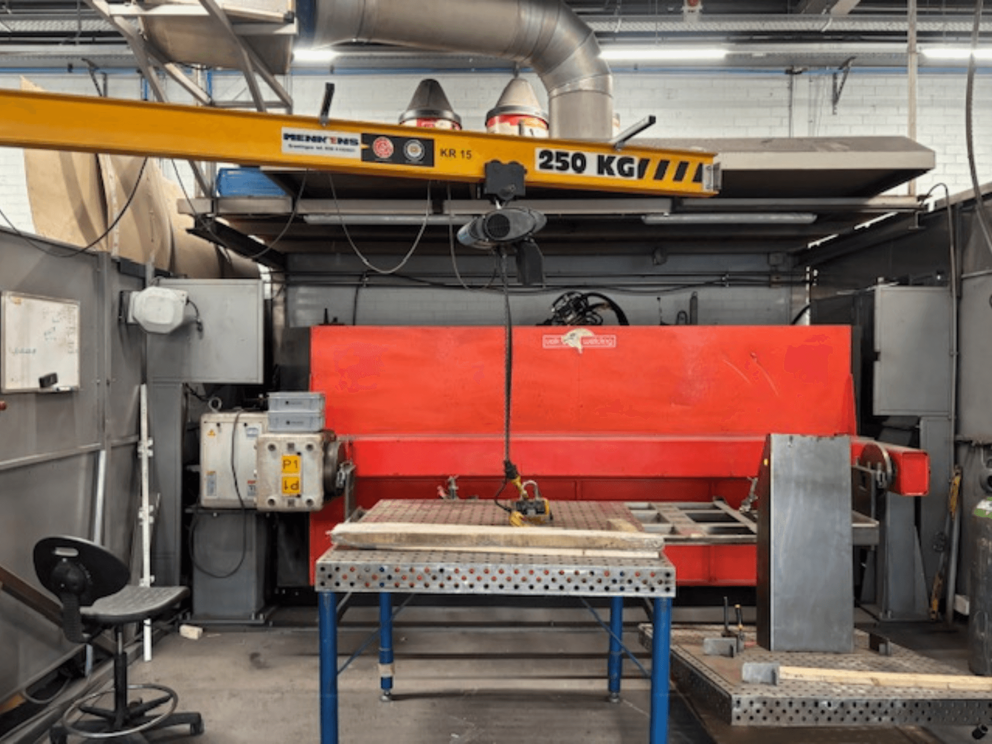 Atelier industriel avec une presse hydraulique rouge, un pont roulant étiqueté "KR 15" et une table de travail en métal, vue latérale.