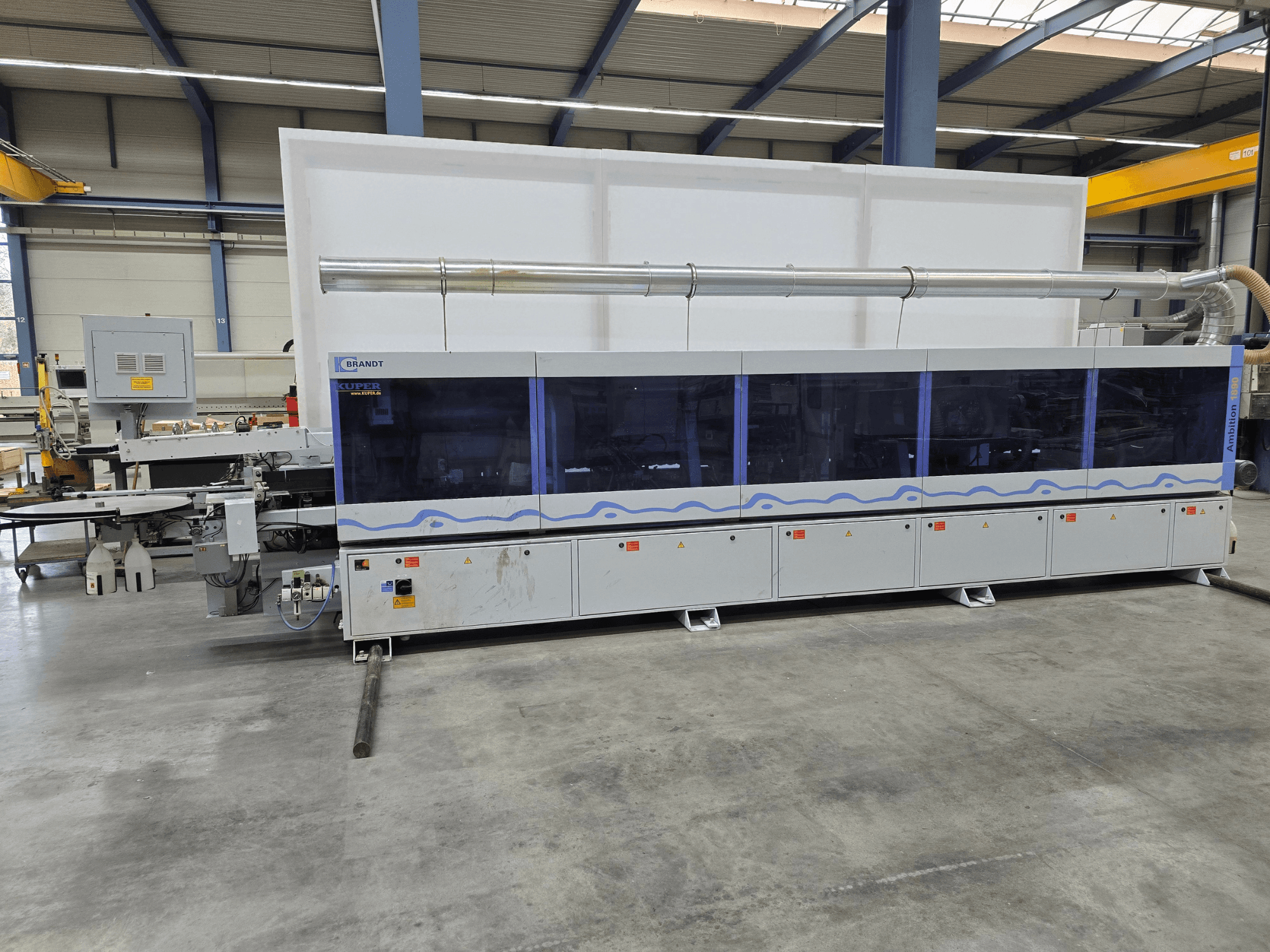 Machine Brandt KUPER dans une usine, vue de face avec panneaux transparents, comportant des boutons de commande et des avertissements de sécurité.