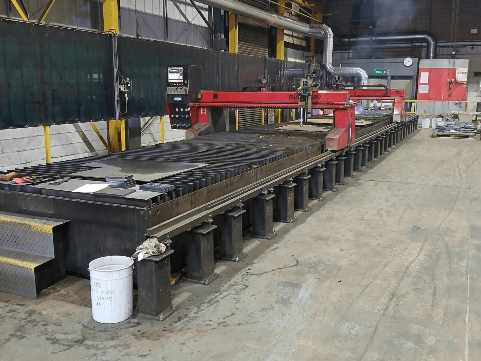Machine à découper RUIDA 300 rouge, vue de côté, avec une tôle sur une table en acier, située dans un espace de travail industriel.
