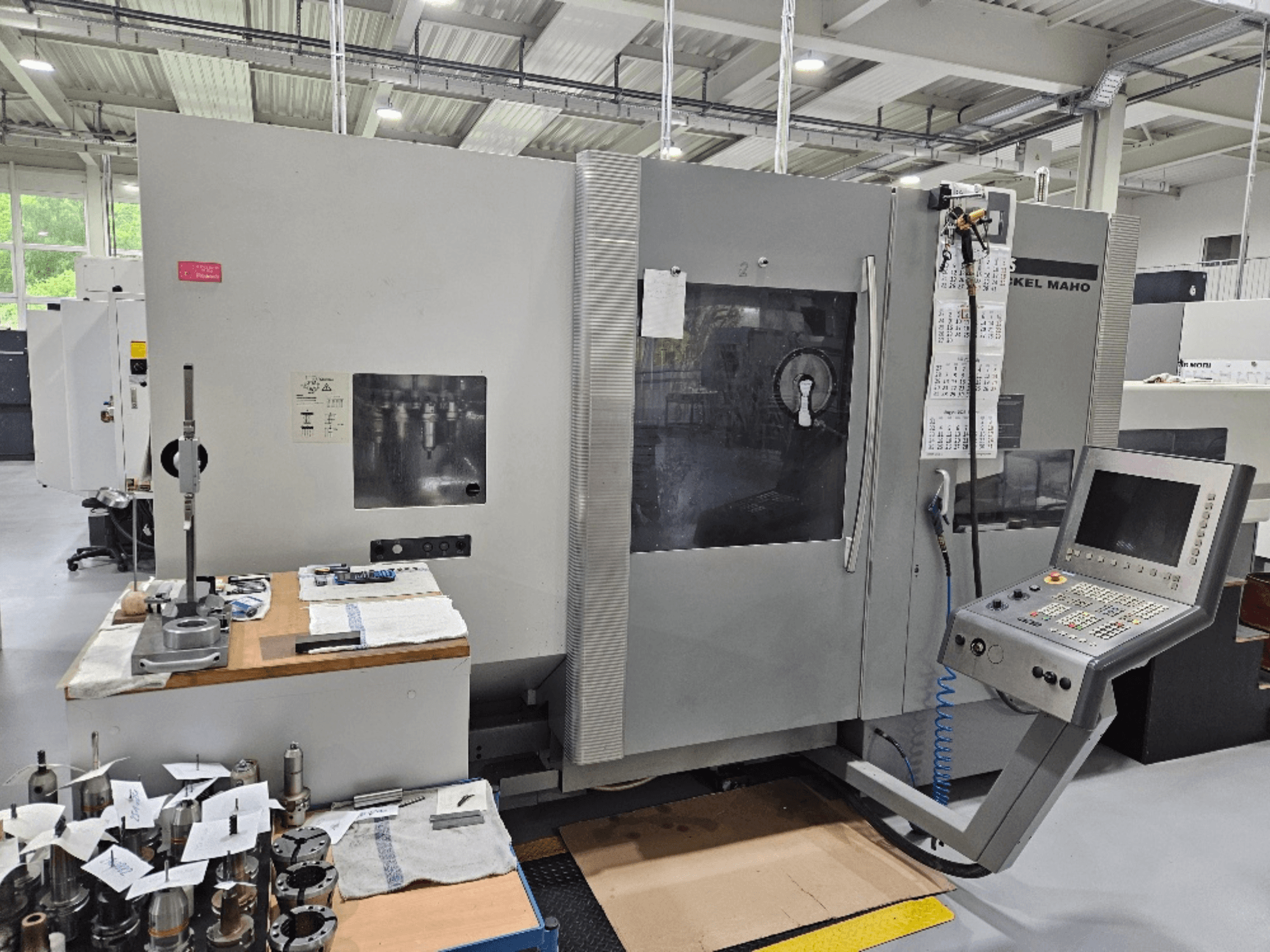 Vue de face d'une machine DMG Mori DMU 70 avec panneau de commande, outils sur une table en bois et diverses pièces à proximité.