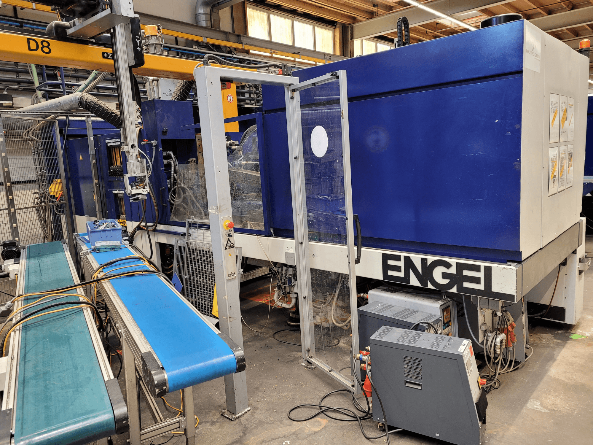 Machine ENGEL bleue avec une porte de sécurité transparente, vue de côté, comportant un panneau de commande, un tapis roulant et des connexions électriques visibles.