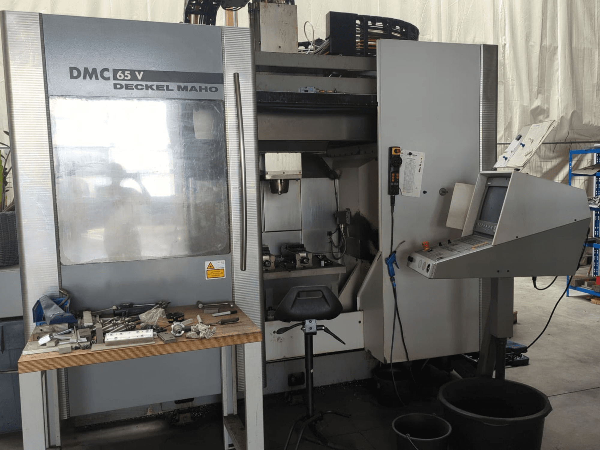 Vue de face d'un centre d'usinage vertical DECKEL MAHO DMC 65 V avec son panneau de commande, ses outils posés sur une table et la configuration de son espace de travail.