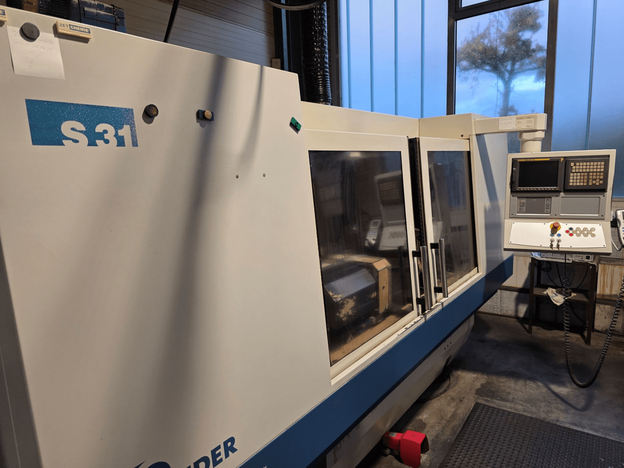 Machine à meuler CNC S31, vue de face, panneau de commande avec écran et boutons, grande porte vitrée, environnement industriel.