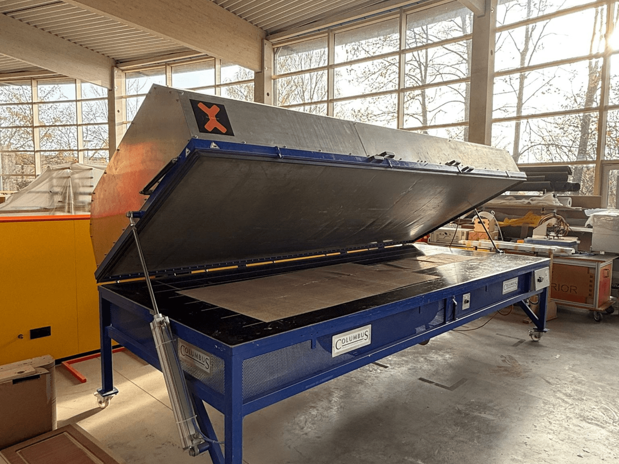 Presse à vide Columbus dans un atelier, vue de face avec le grand couvercle angulaire ouvert, montrant une surface plane et des machines détaillées.