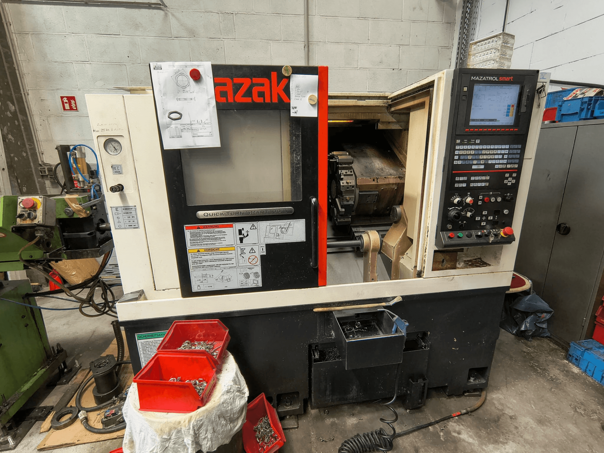 Mazak Quick Turn Smart 100 S, vue de profil montrant le panneau de commande, le bac à liquide de refroidissement et les bacs de rangement contenant des vis.
