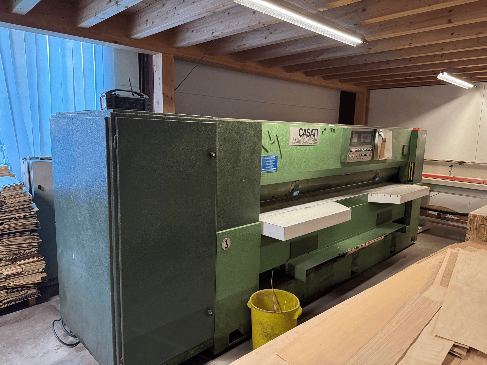 Machine à travailler le bois CASATI verte dans un atelier, vue de face, avec panneau de commande et plans de travail blancs, entourée de piles de bois.