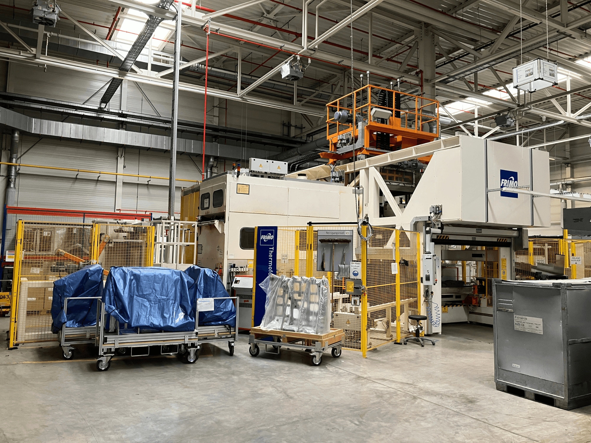Machine de transformation du plastique FRIMO FTL 80/200 Fullflex dans une usine, vue de face avec des bâches bleues et des barrières de sécurité visibles.