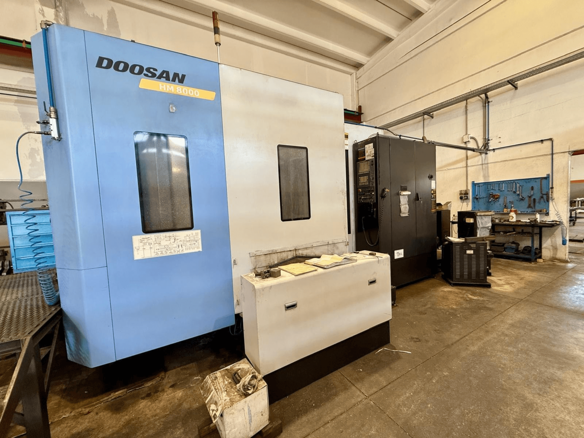 Centre d'usinage horizontal DOOSAN HM 8000 de couleur bleue, vue de face, montrant le panneau de commande et les équipements d'atelier situés à proximité.