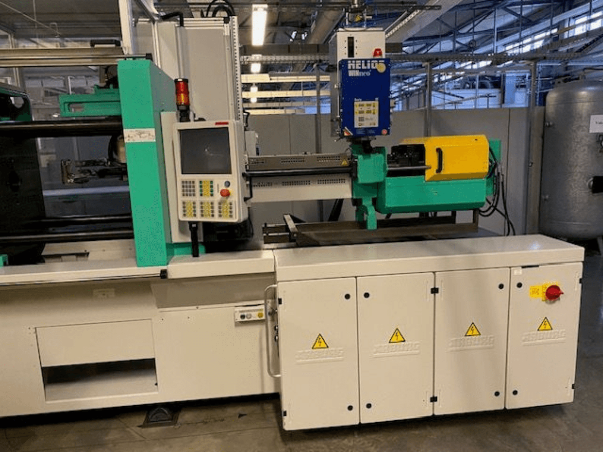 Machine industrielle de moulage par injection, modèle HELIO, vue de face, comprenant le panneau de commande, les protections de sécurité et le bouton d'arrêt d'urgence.