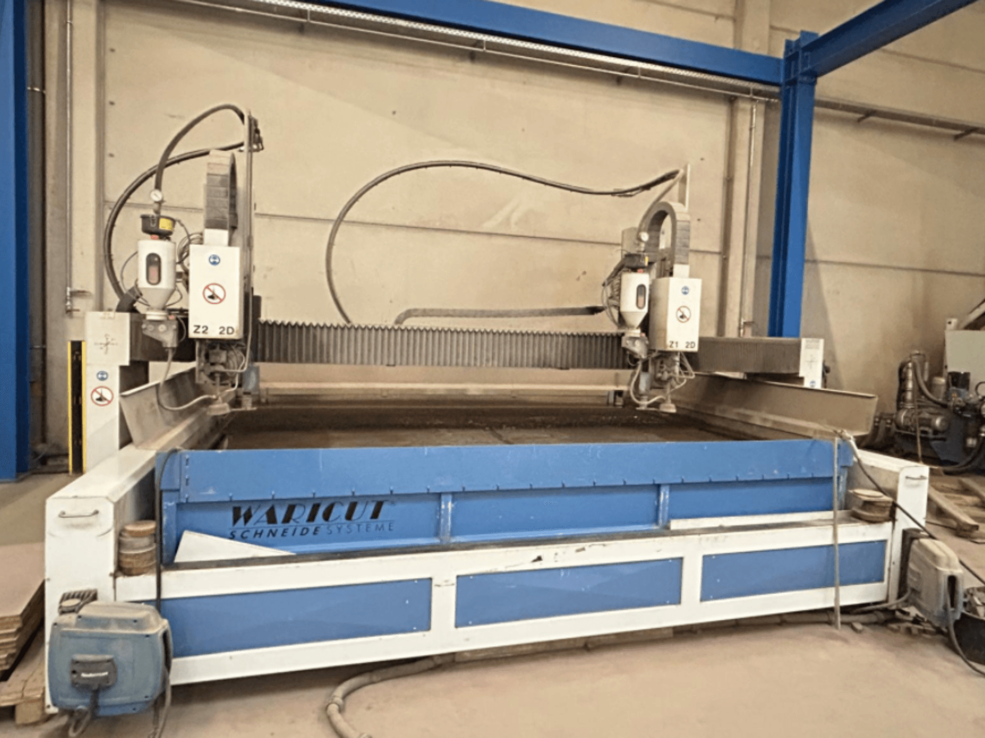 Machine de découpe au jet d'eau de Waricut, vue de face ; elle est équipée de deux têtes de découpe et d'une surface de travail bleue dans un atelier.