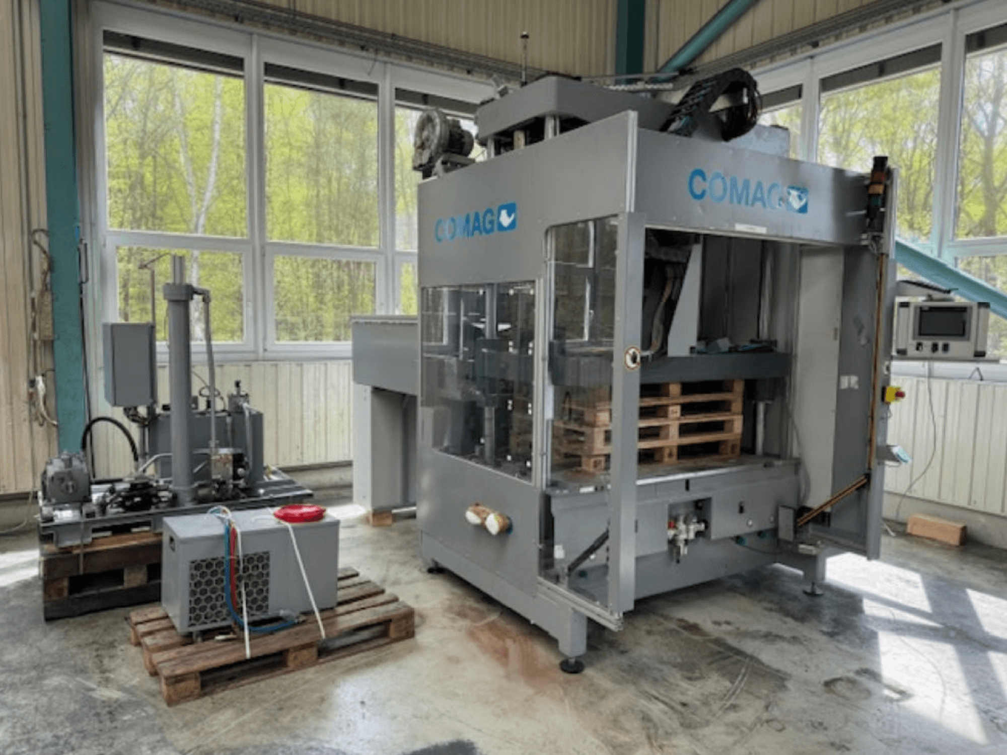 Machine d'emballage de palettes COMAG dans un environnement industriel, vue de face ; les palettes et les composants hydrauliques sont visibles.