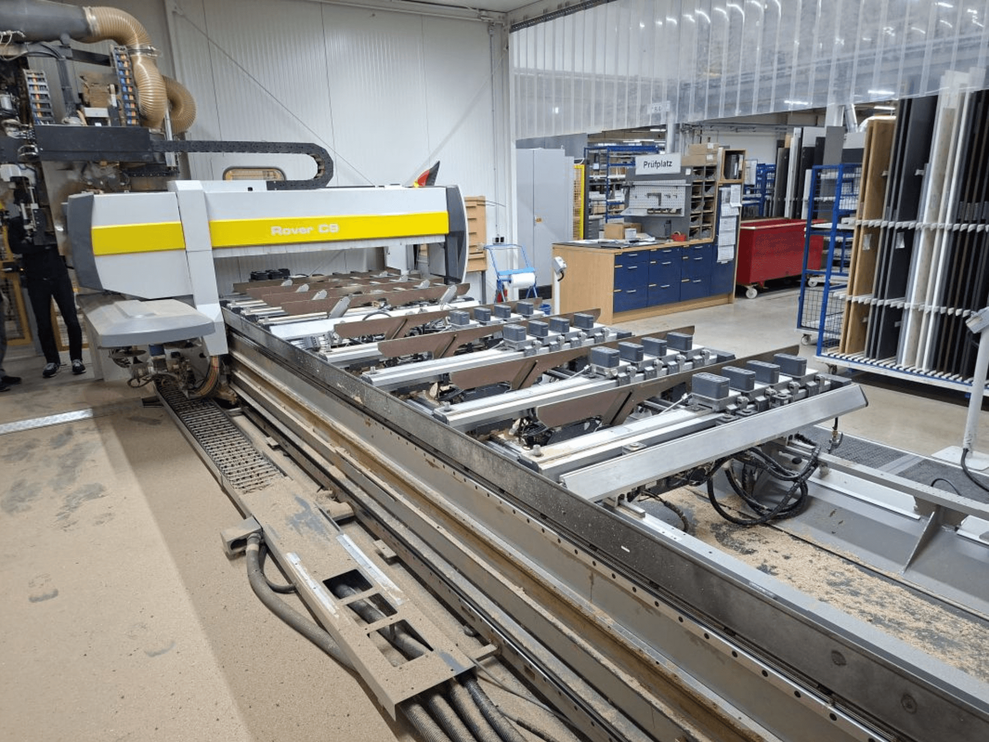 Machine CNC Rover C9 dans un atelier ; la vue de face montre les outils de coupe et le système de convoyage, avec une accumulation de poussière sur le sol.