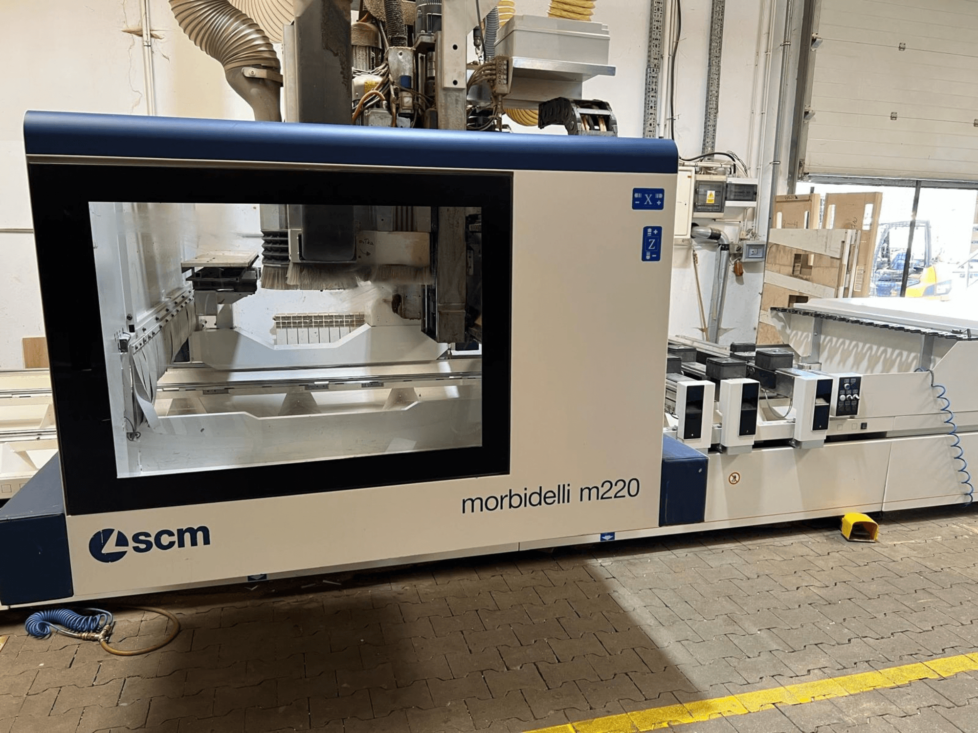 Vue de face du centre d'usinage CNC SCM Morbidelli M220, mettant en évidence une grande vitre, le panneau de commande et la zone de travail.