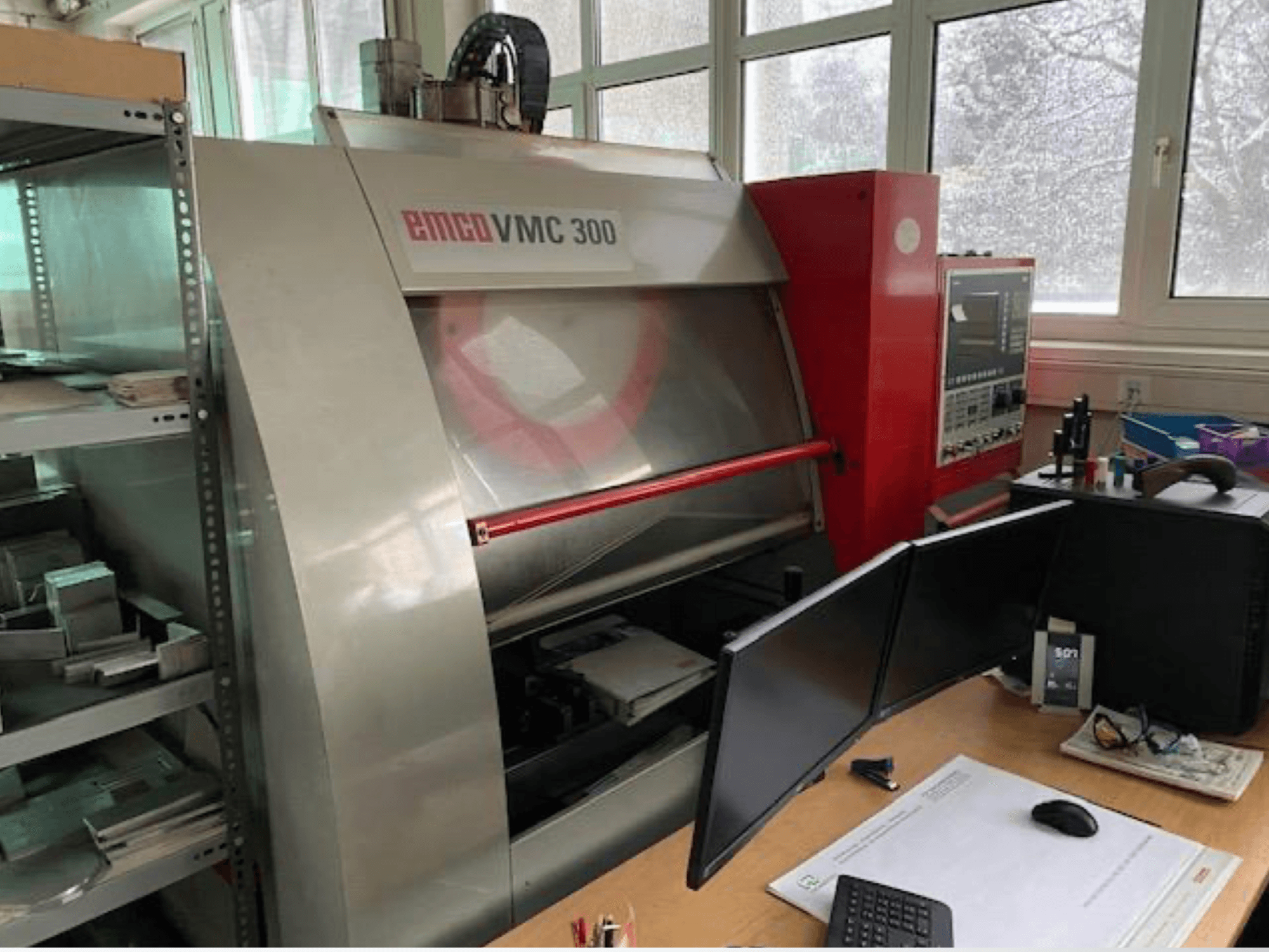 Centre d'usinage vertical EMCO VMC 300 dans un atelier, vue de profil montrant le panneau de commande et le poste de travail adjacent équipé de deux écrans.