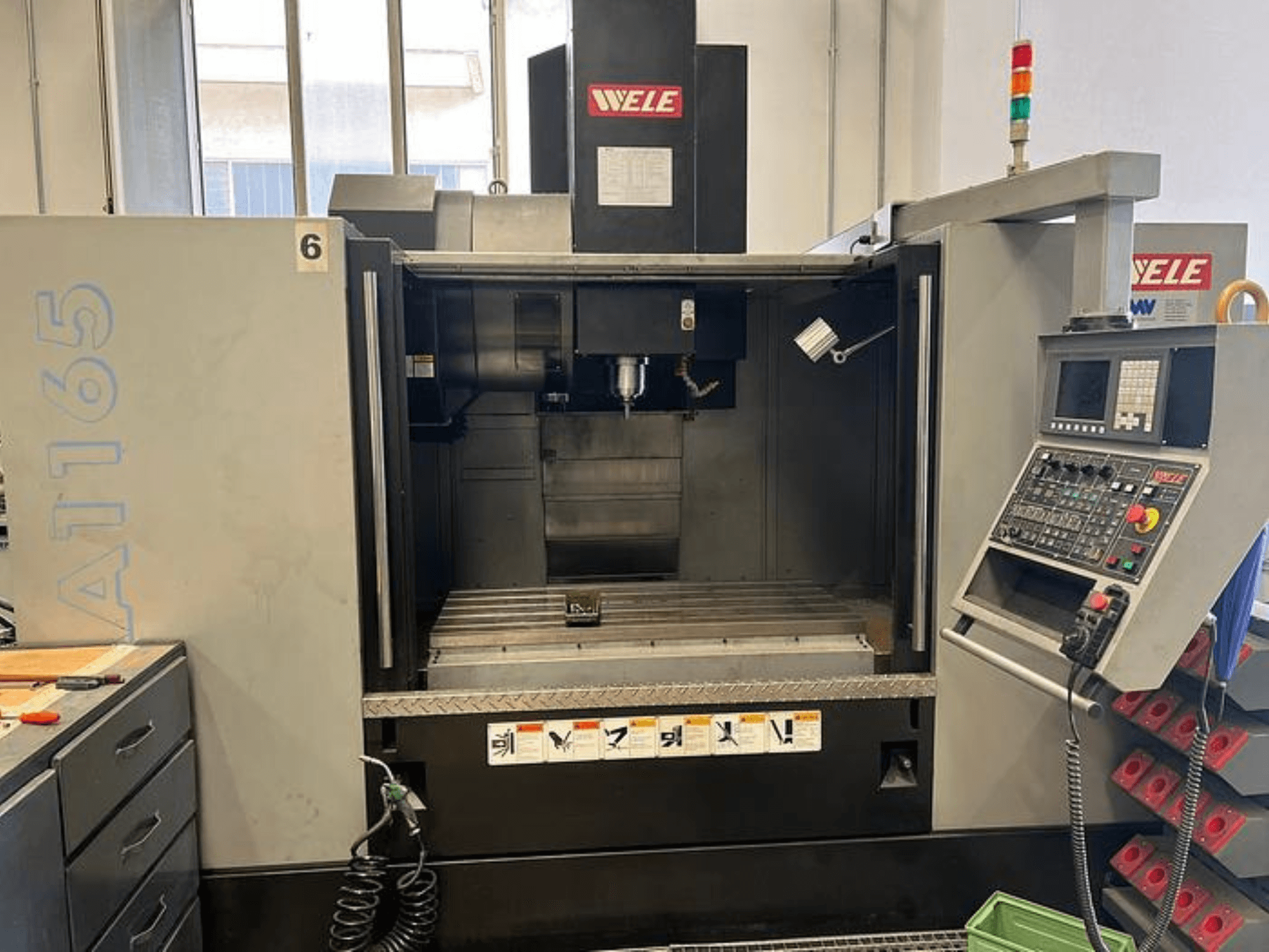 Centre d'usinage CNC WELE A165, vue de face, avec le panneau de commande, le porte-outil et la table de travail visibles. Environnement industriel.