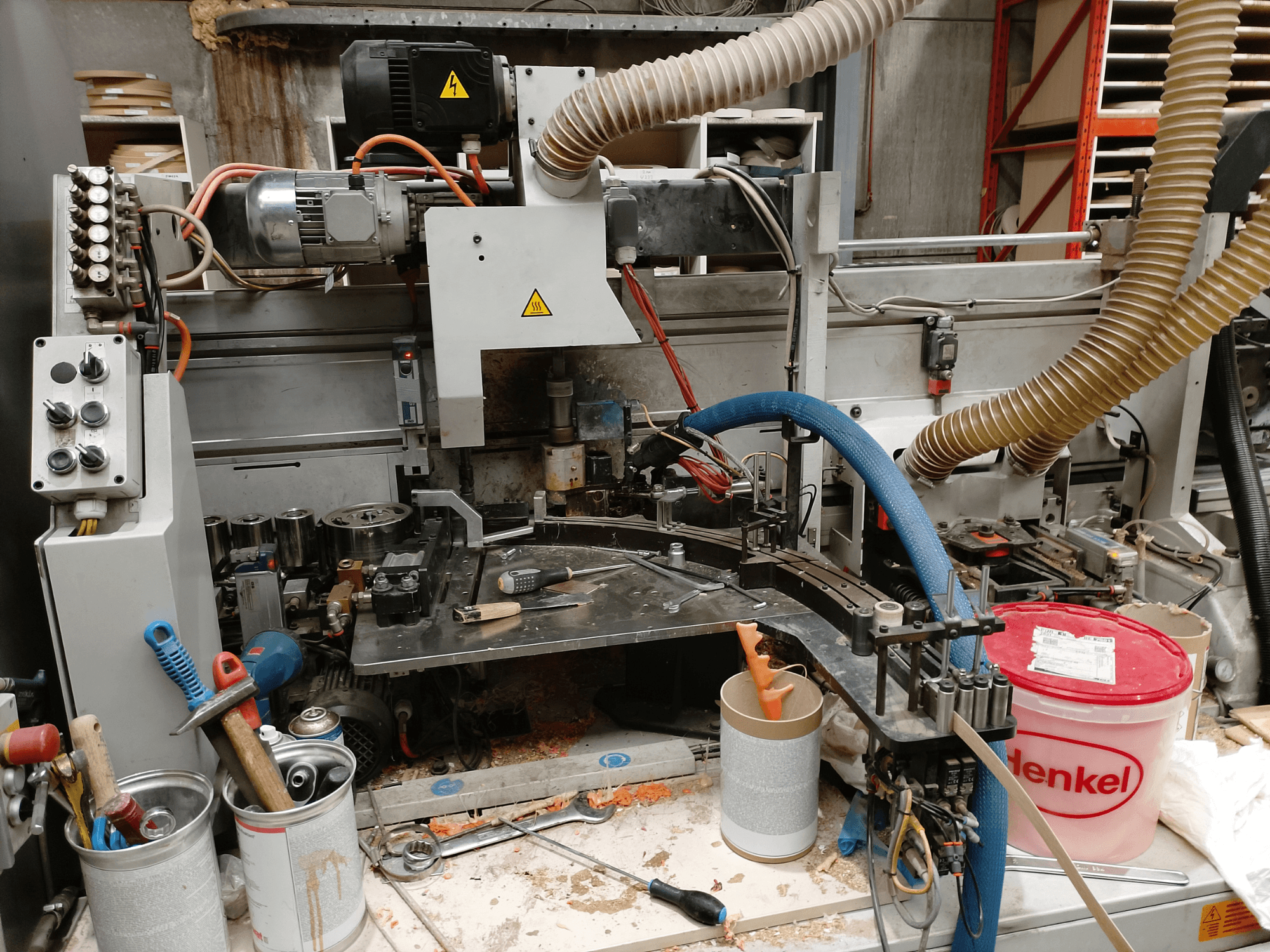 Plaqueuse de chants BIESSE Edge Stram B1 en vue de face, présentant les machines, les tuyaux et les outils de travail au milieu des copeaux de bois.