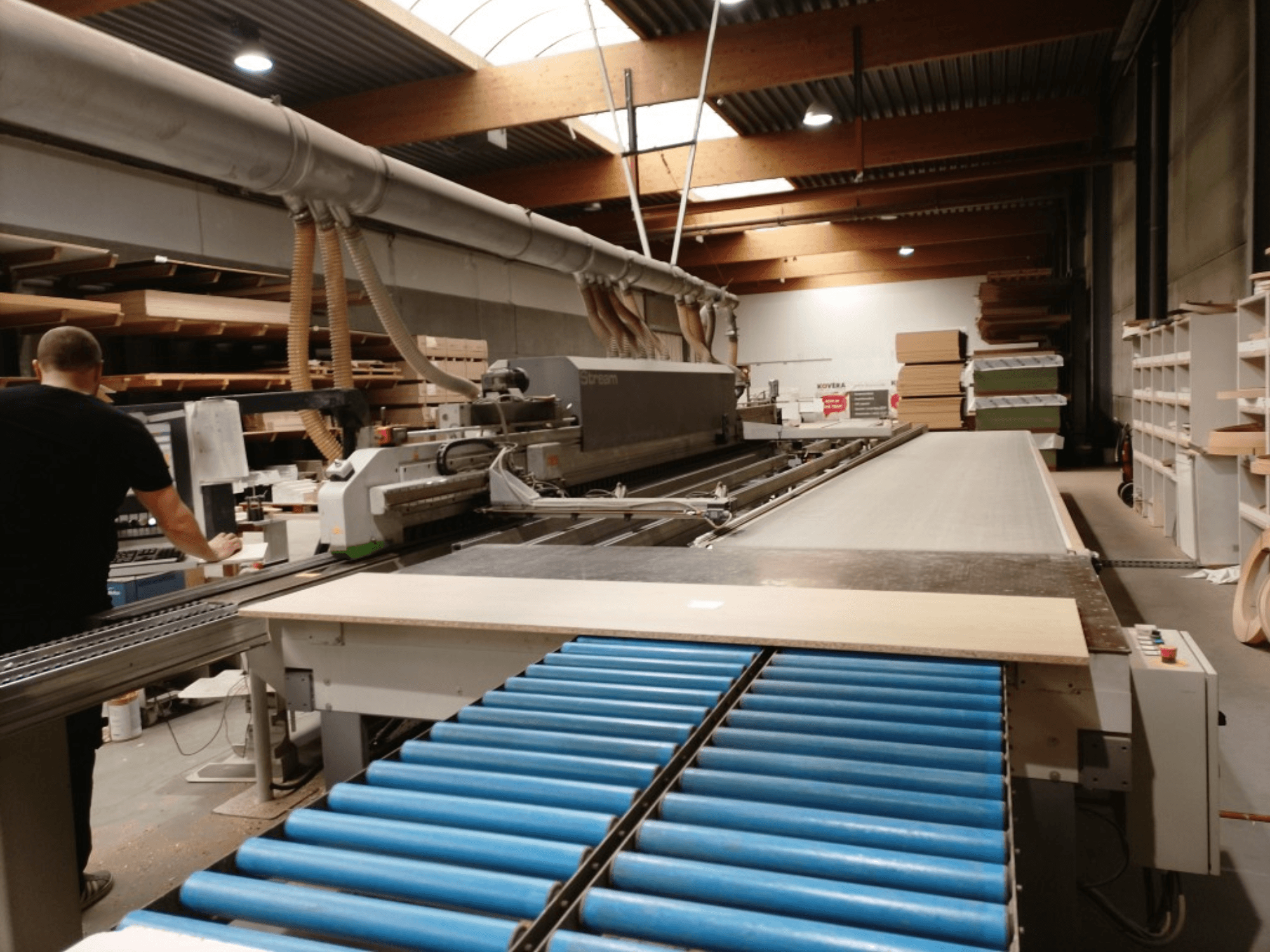 Plaqueuse de chants BIESSE Edge Stream B1, vue latérale montrant l'opérateur, les rouleaux bleus et le panneau de bois sur le convoyeur.