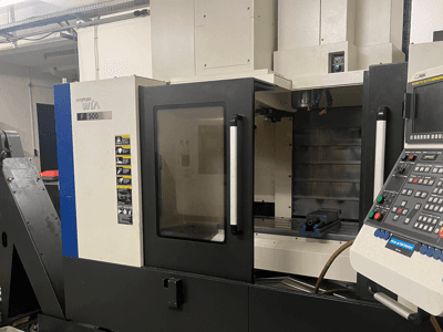 Centre d'usinage vertical Hyundai Wia F500, vue de face, montrant le panneau de commande, la zone de travail équipée d'un étau bleu et les étiquettes d'avertissement.