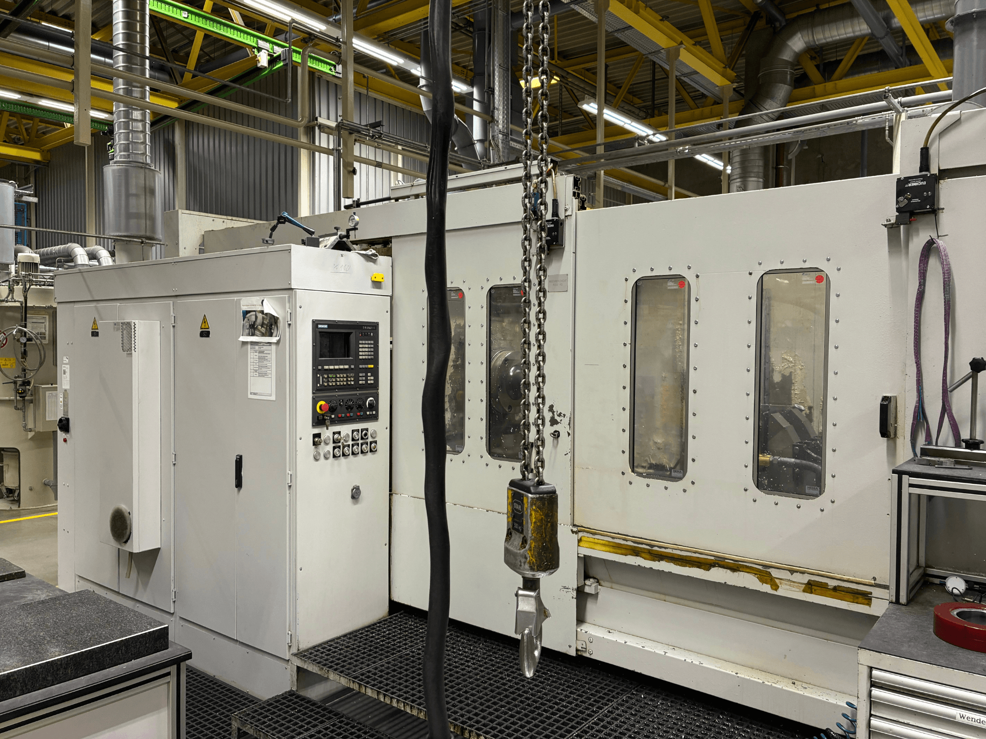 Machine industrielle à commande numérique avec panneau de contrôle, vue de côté ; on y voit des fenêtres, des chaînes et des composants métalliques dans une usine.