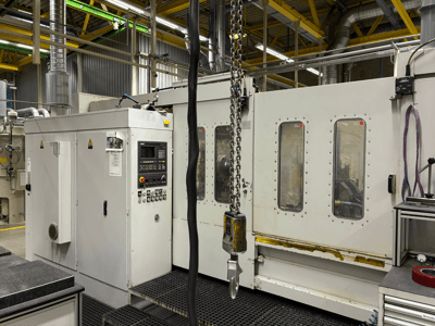 Machine industrielle à commande numérique avec panneau de contrôle, vue de côté ; on y voit des fenêtres, des chaînes et des composants métalliques dans une usine.